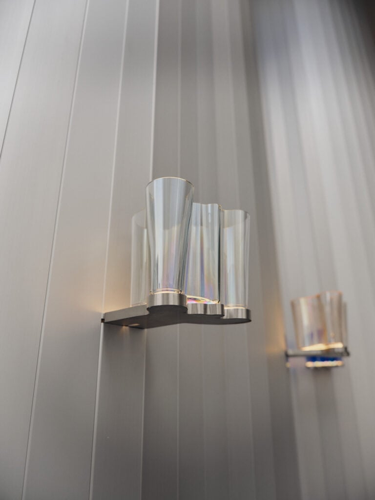 Wall-mounted light fixture with a fluted glass shade attached to a metallic base on a vertically paneled wall; another similar fixture is visible in the background, reflecting the contemporary style seen at Milan Design Week 2026.