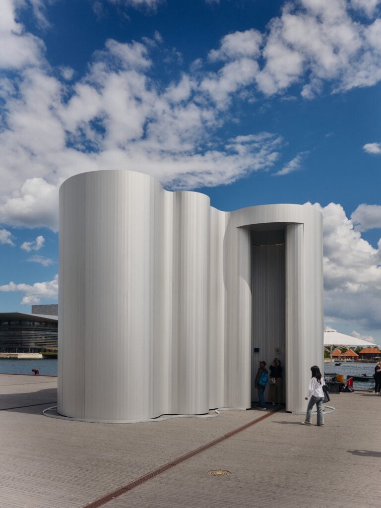 A modern, curved white pavilion stands outdoors on a paved area at Milan Design Week 2026, with people walking nearby under a partly cloudy sky.