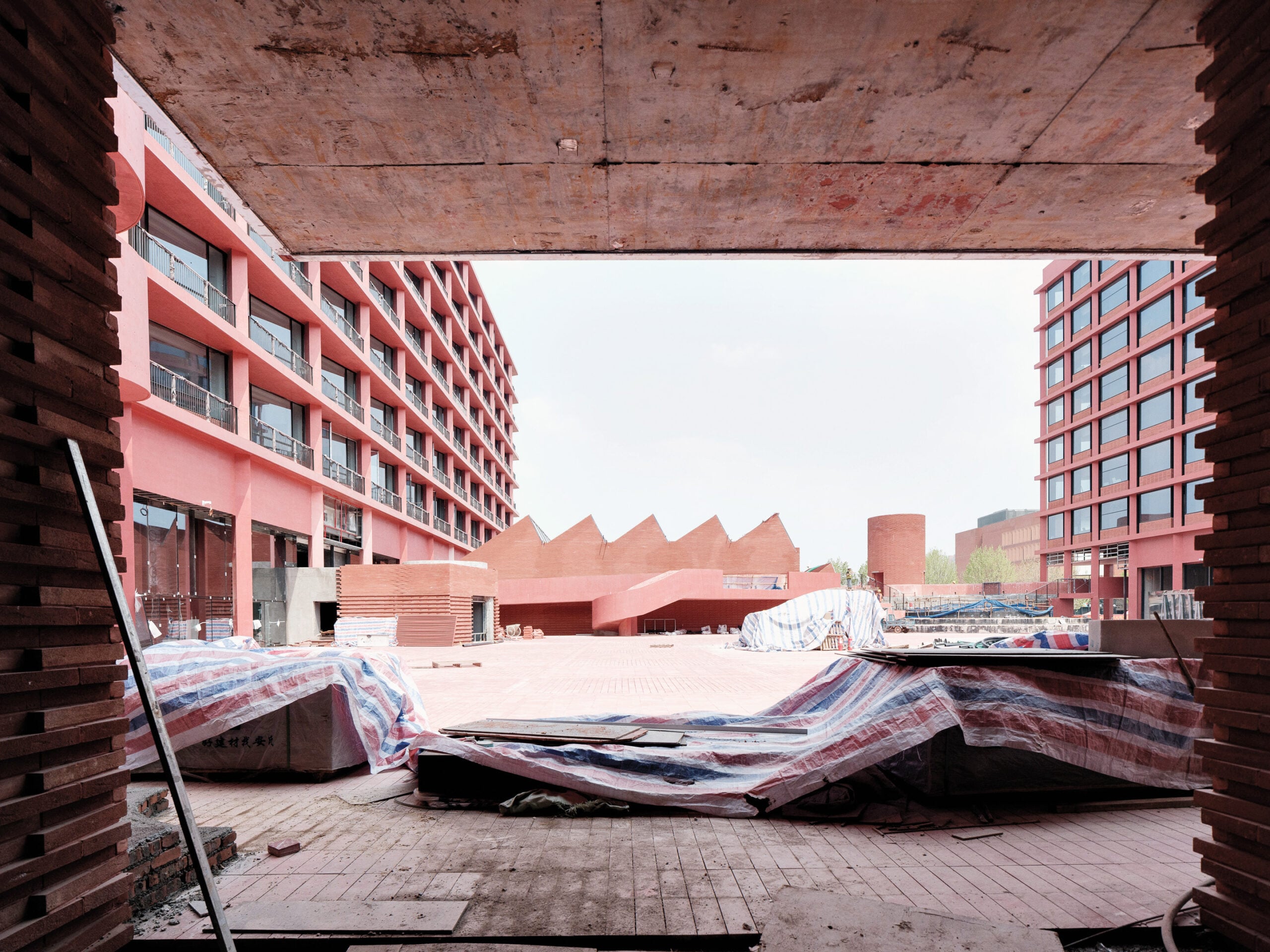 View of a modern red-brick building complex under construction, with materials and scaffolding in the foreground and angular rooflines visible in the courtyard.