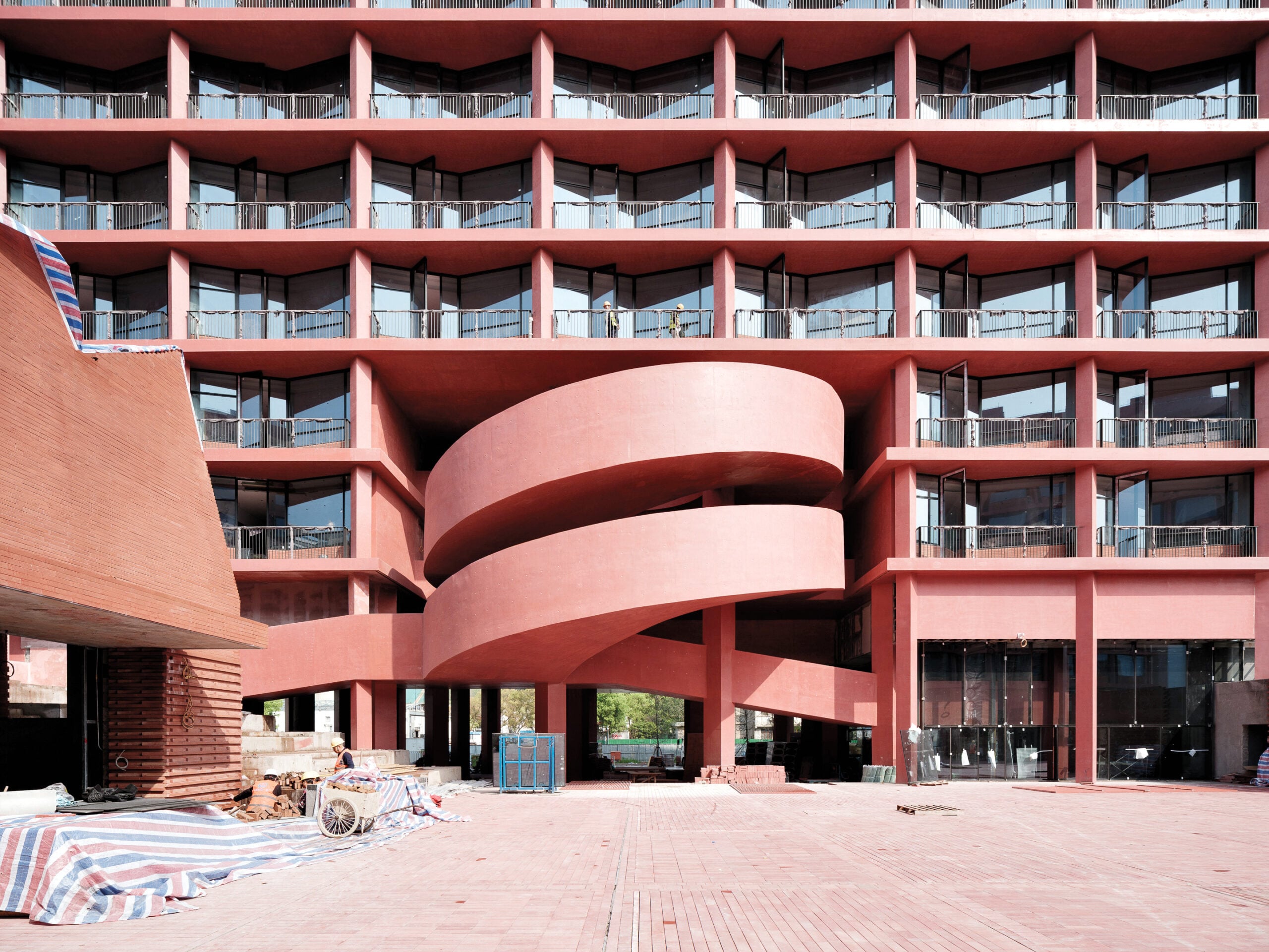 A modern red building facade with multiple balconies and a prominent curved spiral ramp at the entrance, surrounded by construction materials.