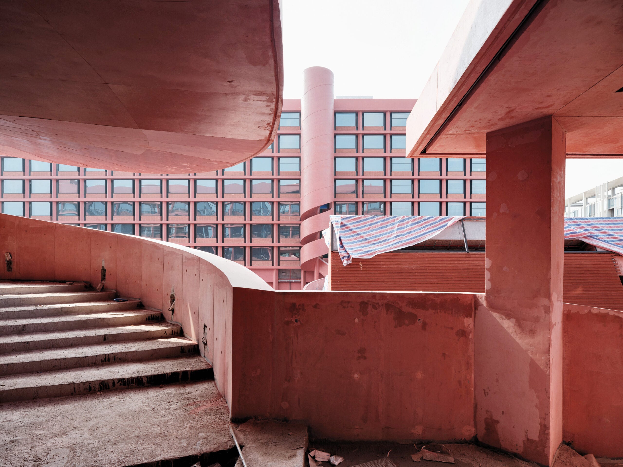 Curved concrete stairs and walkways in an unfinished red building, with a multi-story glass and red panel facade in the background.