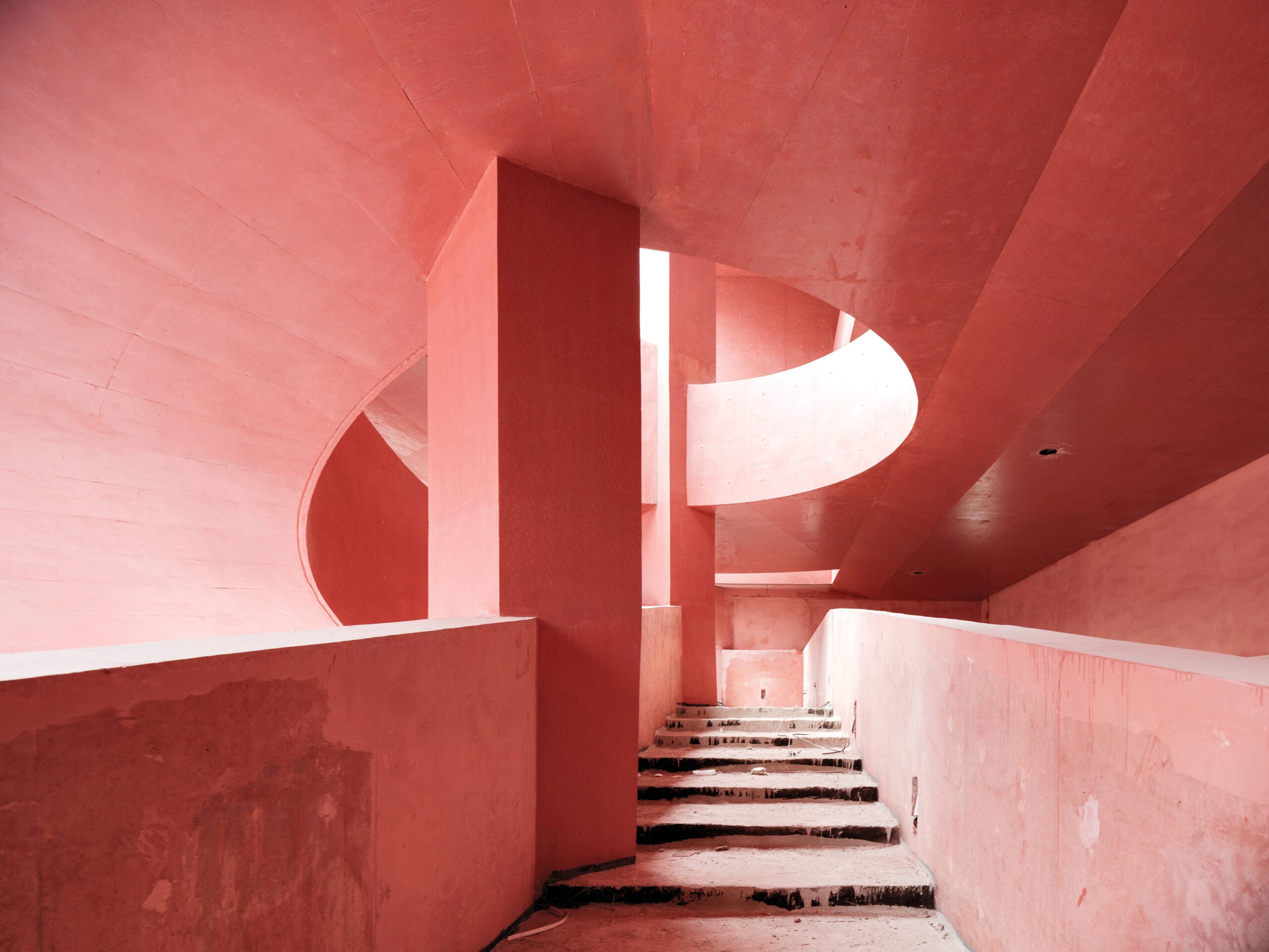 A staircase with unfinished steps and pink concrete walls curves upward beneath a circular skylight in a modern architectural interior.