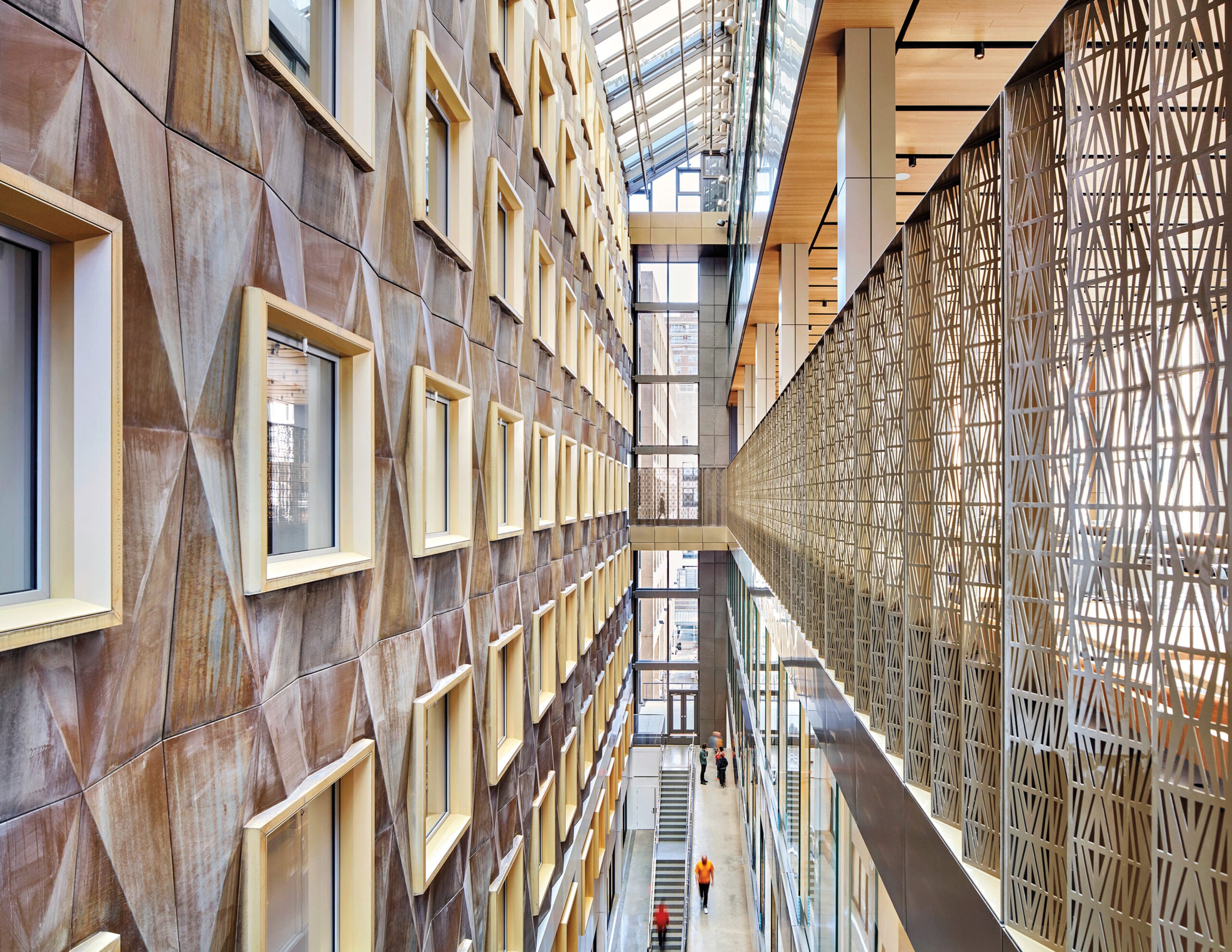 Craft in Architecture: Interior view of a modern building atrium with geometric wall patterns, large windows, a glass ceiling, and people walking along the corridor below.