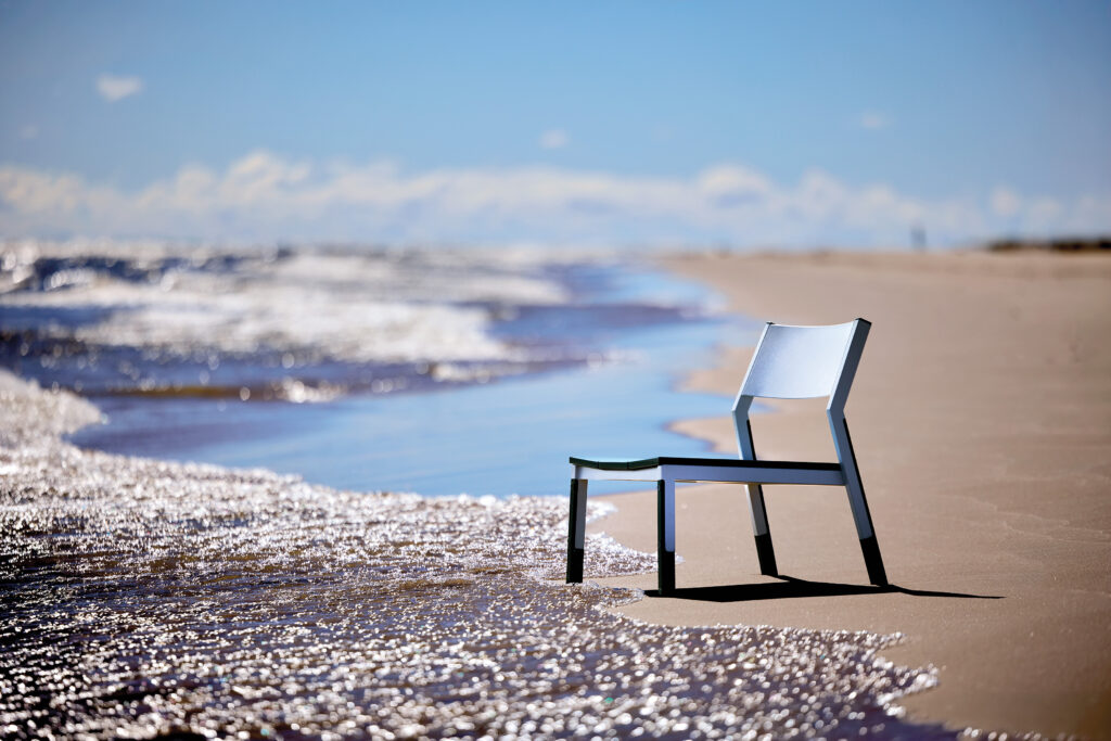A single chair with broken front legs sits on a sandy beach near the shoreline, partially surrounded by incoming waves under a clear sky.