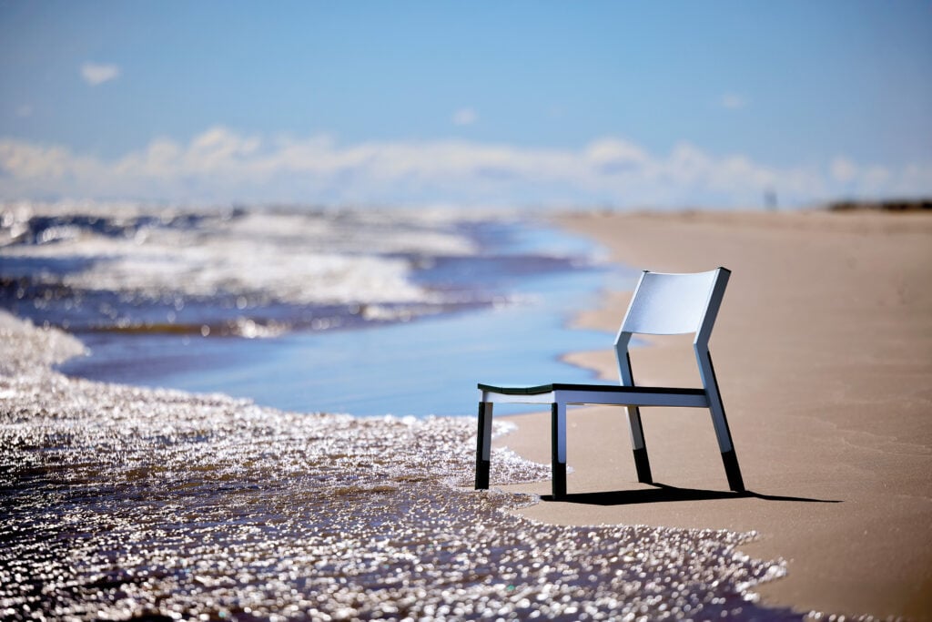 A metal chair with two missing legs is partially submerged at the edge of the ocean on a sandy beach under a clear blue sky, capturing an image METROPOLISLikes for its urban decay meeting serene nature.