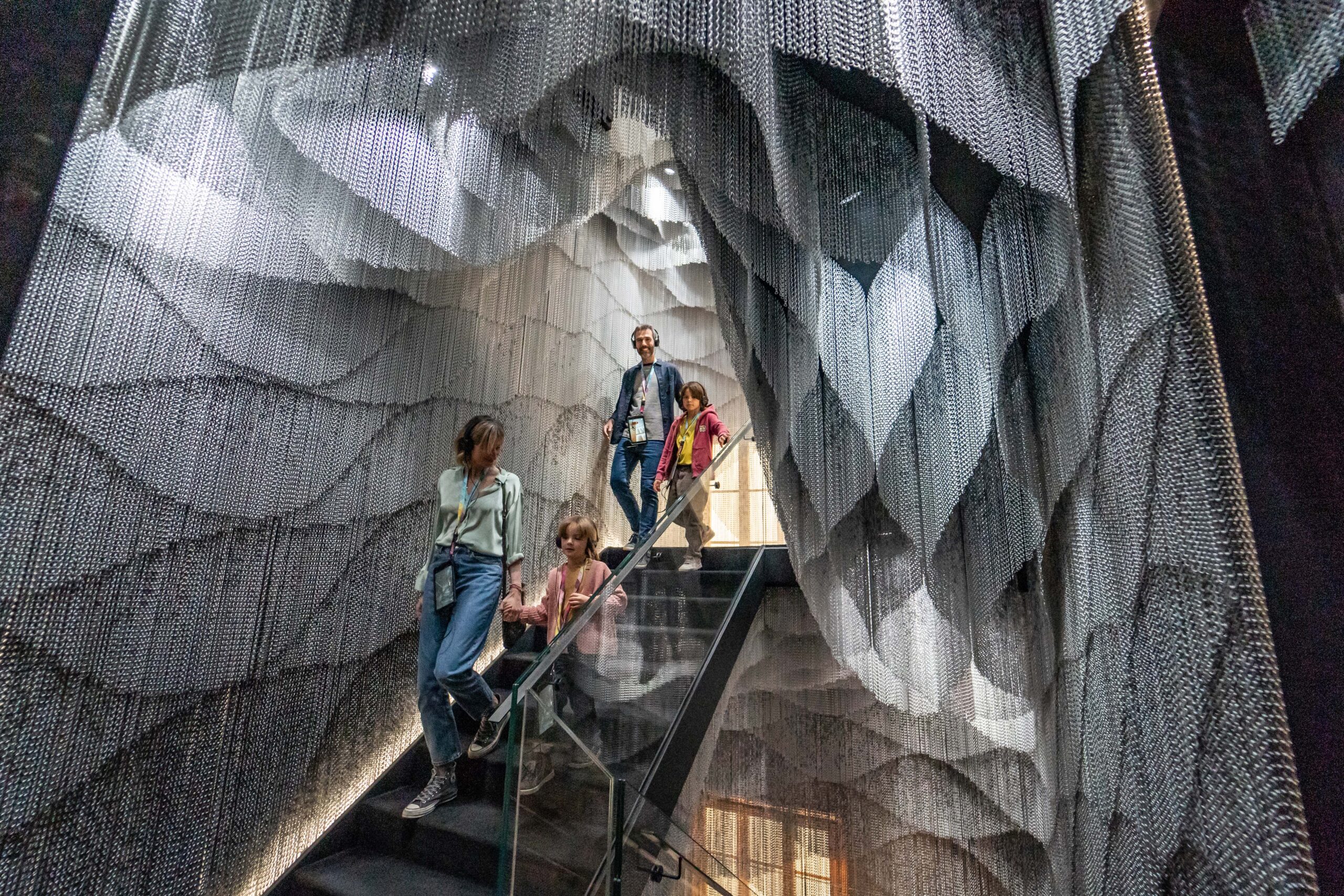 Four people descend a glass staircase, surrounded by intricate hanging metal chains creating a dramatic, textured architectural installation.
