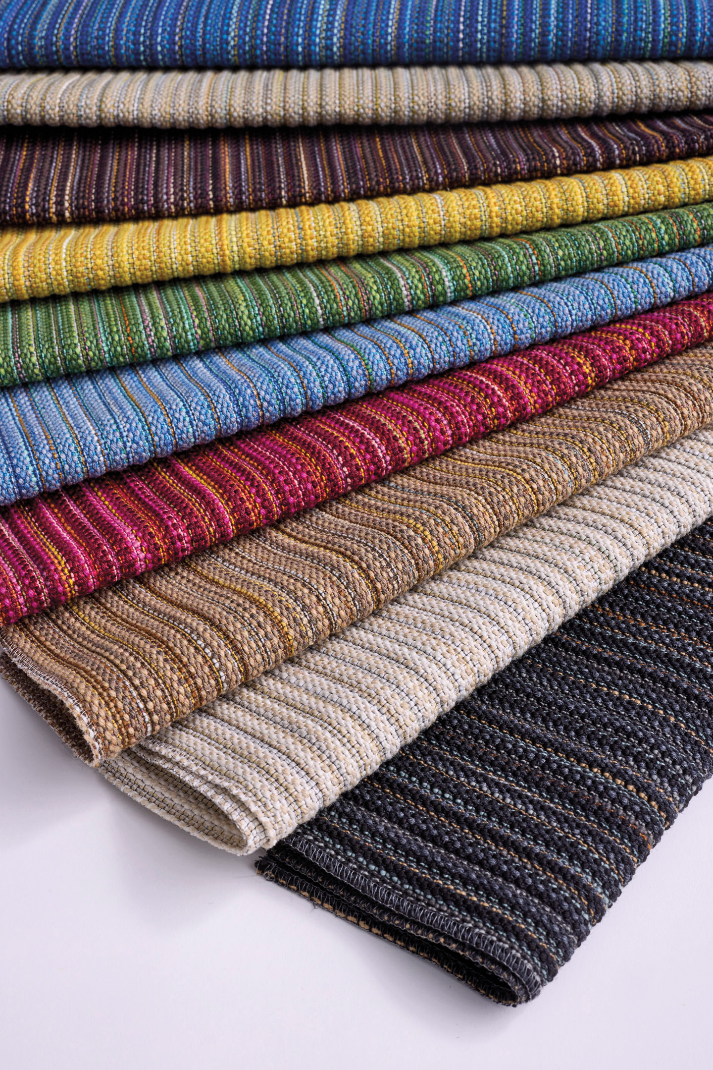 Stacks of folded, striped fabric by Dorothy Cosonas for Luum in various colors including blue, yellow, red, brown, beige, and black, arranged in a neat overlapping pattern.
