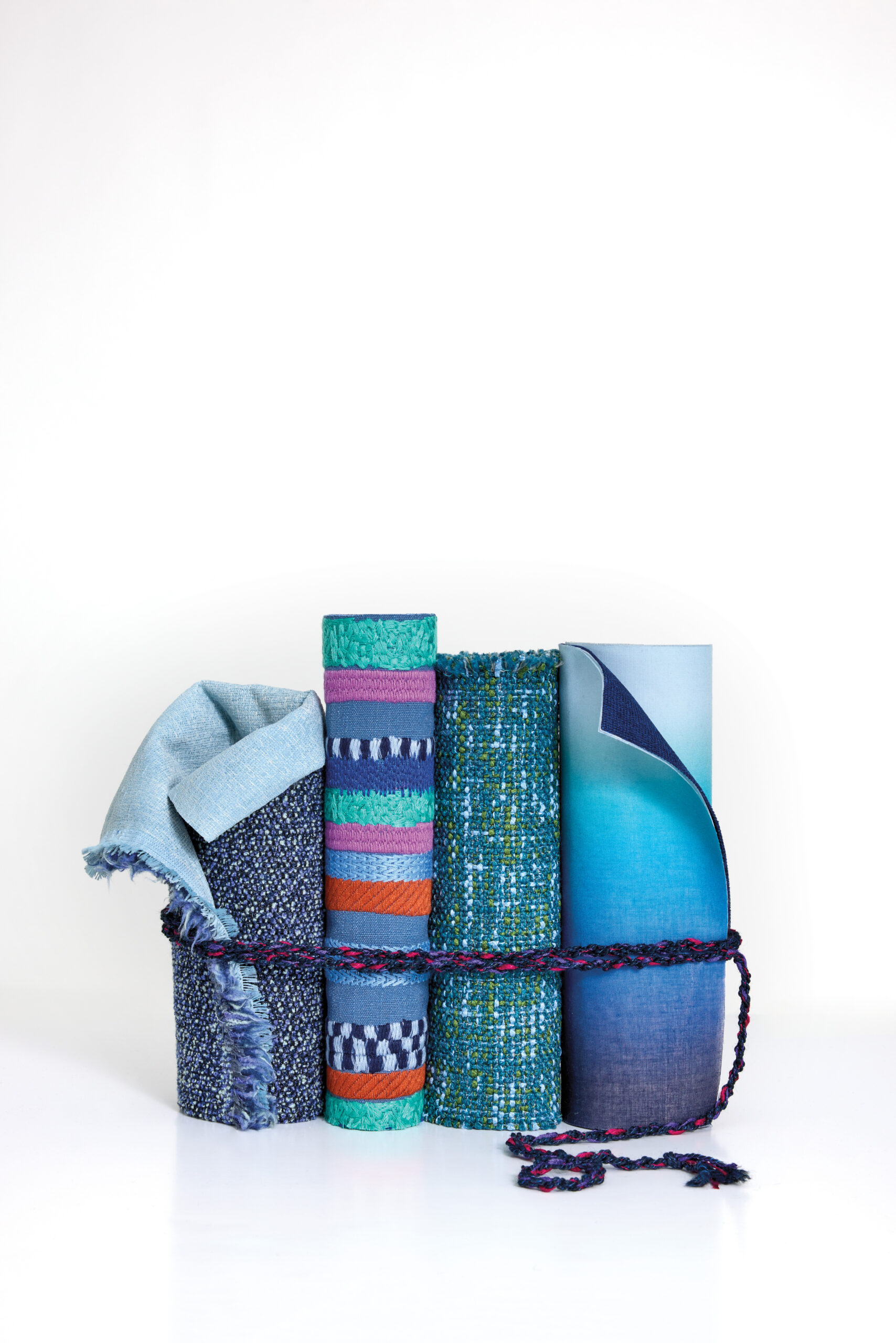 Four rolled textiles in shades of blue and teal, some with patterns, are grouped together and tied with a multicolored braided cord against a white background.