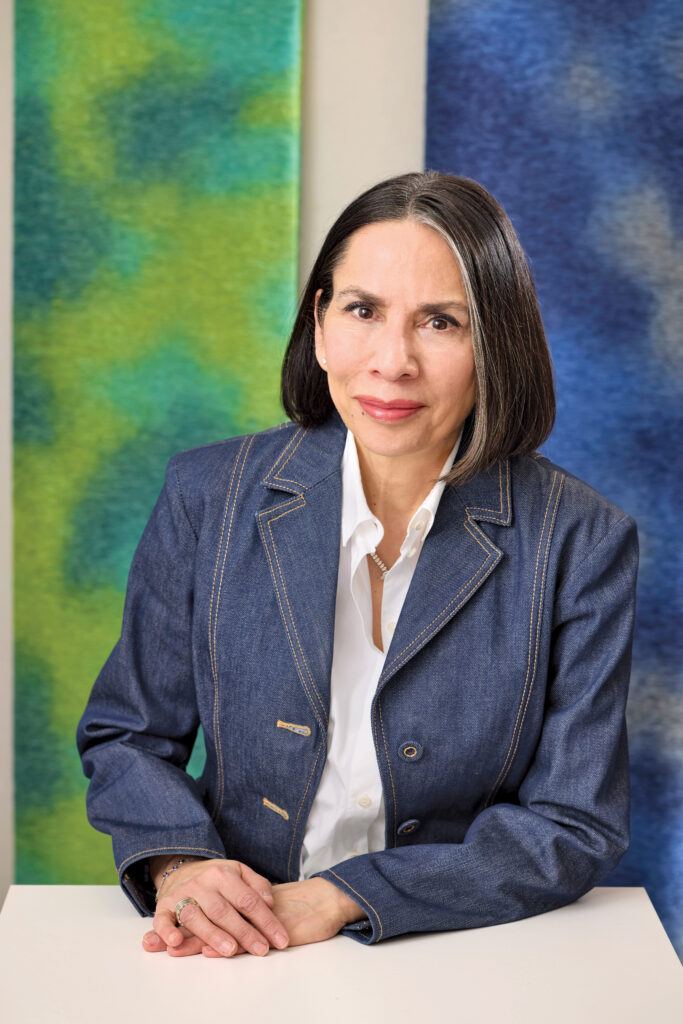 A woman with straight dark hair, Dorothy Cosonas, wearing a denim blazer and white shirt, sits at a table in front of green and blue abstract backgrounds.