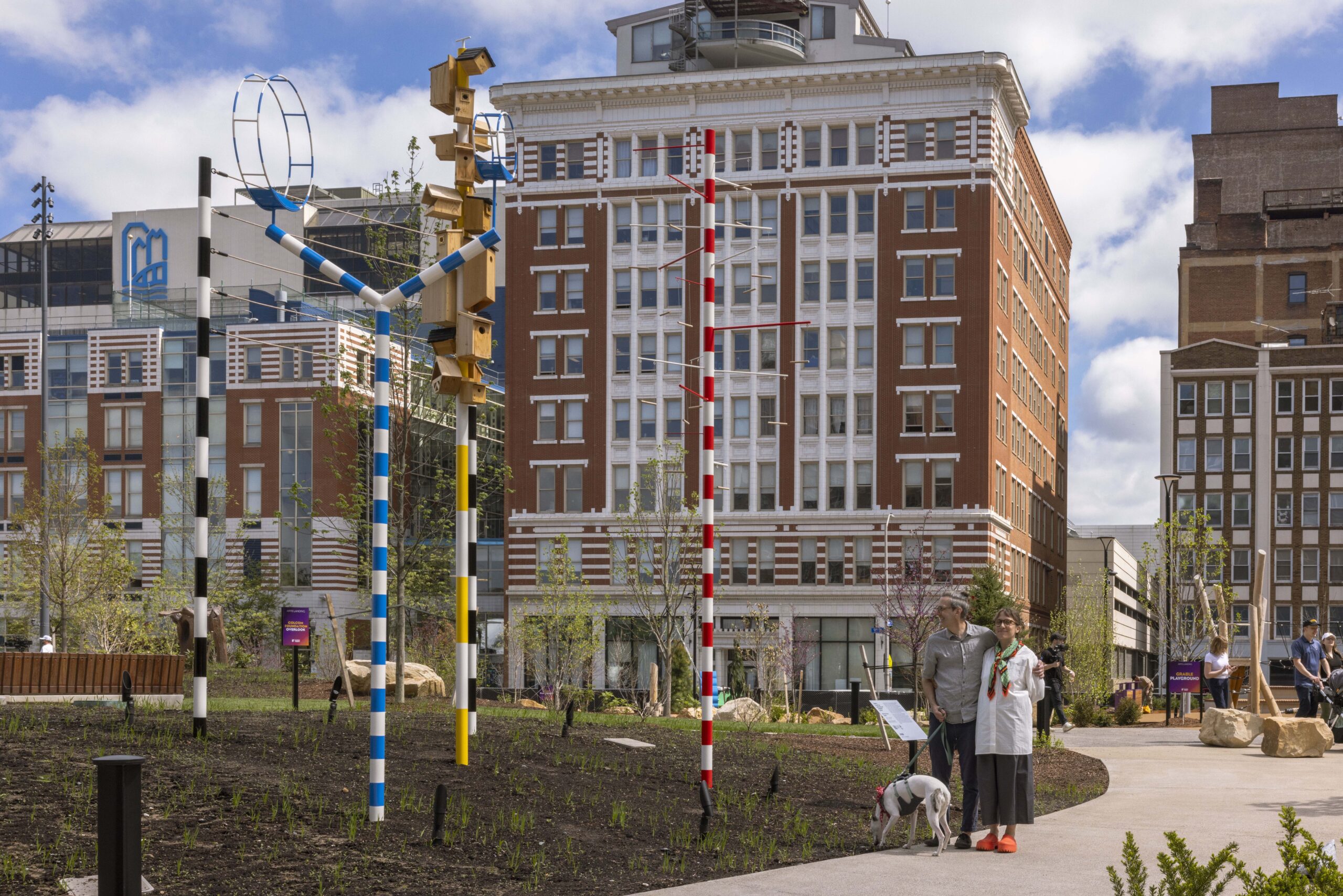 People walk on a path next to colorful striped sculptures in an urban park designed by Field Operations, with tall buildings rising in the background under a partly cloudy sky.