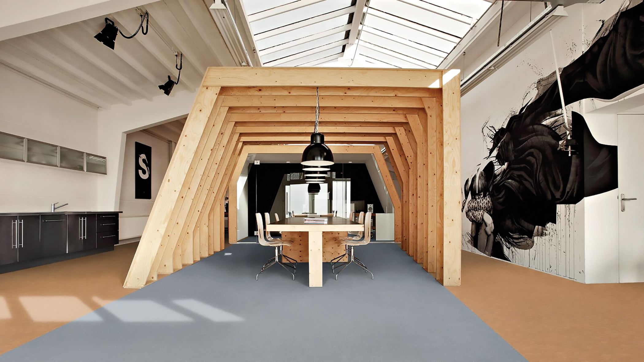 Modern office space features a wooden geometric meeting pod with a long table and chairs; skylights above and a black-and-white mural decorate the wall to the right.