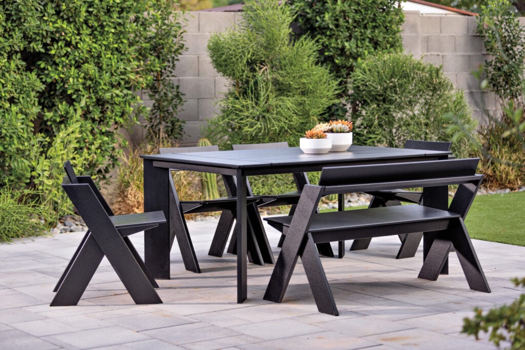 A modern black outdoor dining set with two benches and two chairs is placed on a stone patio, surrounded by greenery and a concrete block wall. METROPOLISLikes style shines with two white planters filled with succulents on the table.