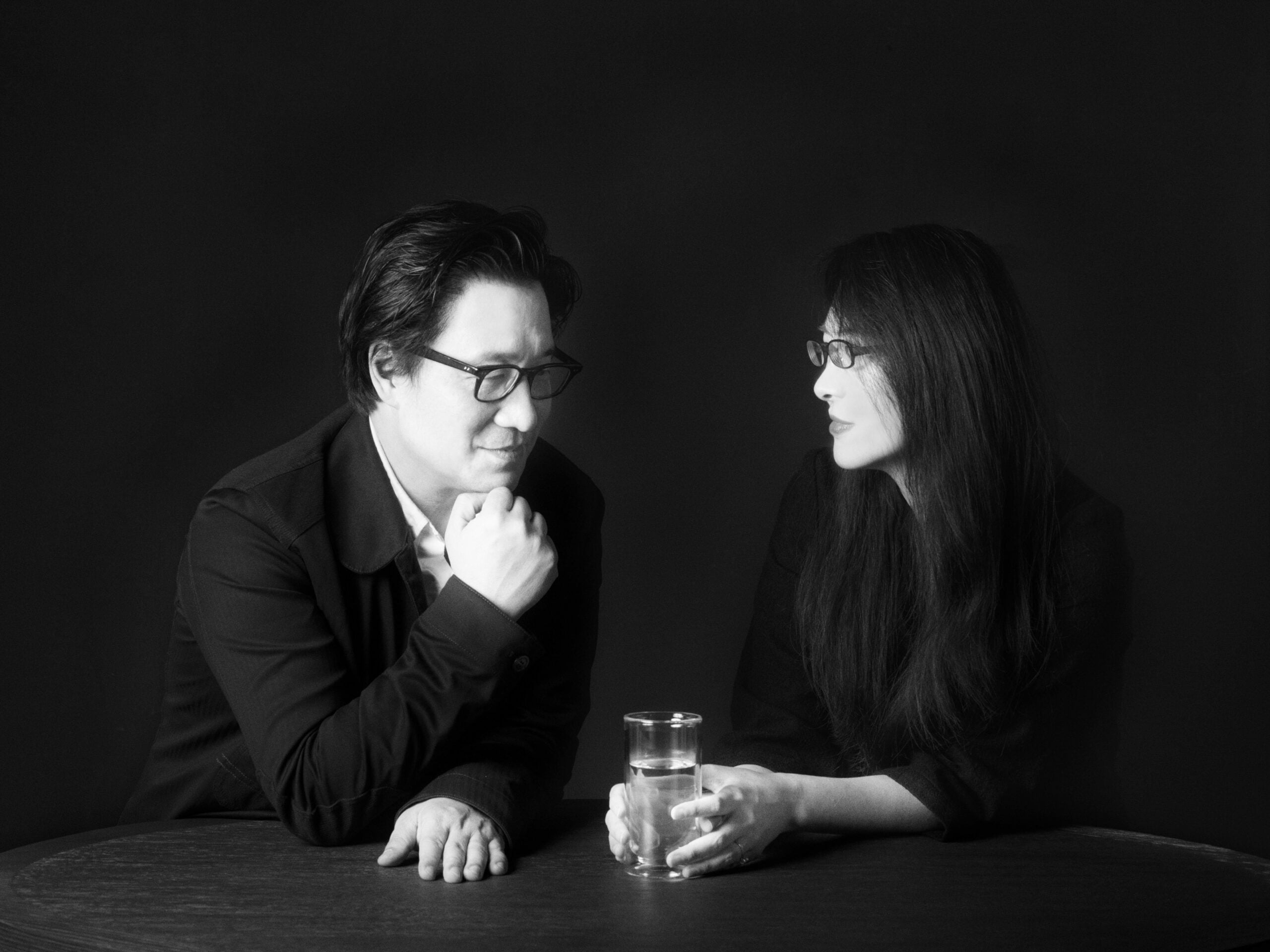 The two founding principals of Neri&Hu sit at a table with a glass of water, facing each other against a dark background. Both wear glasses and dark clothing.