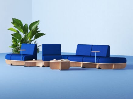 Modular blue and wood seating with built-in tables arranged on a light blue floor, with a potted plant in the background against a light blue wall.