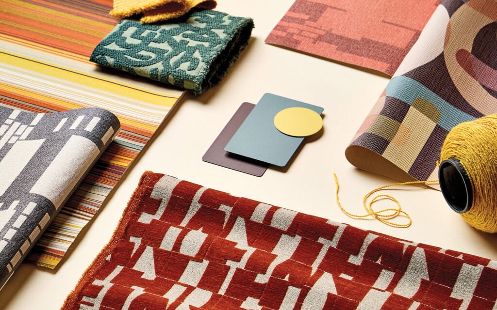 Assorted patterned textiles, color swatches, and a spool of yellow thread are arranged on a beige surface.