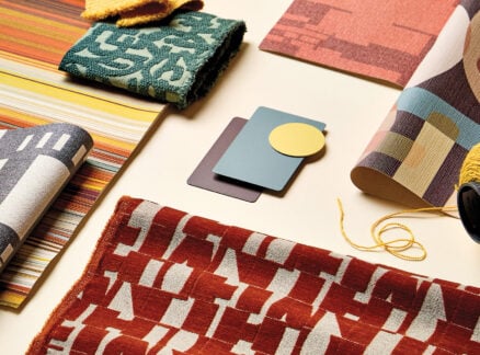 Assorted patterned textiles, color swatches, and a spool of yellow thread are arranged on a beige surface.