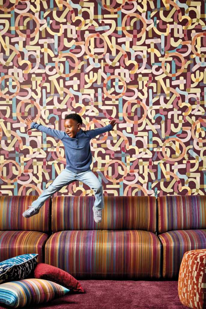 A young boy in a blue shirt and light pants jumps on a striped couch in front of a colorful, geometric-patterned wall, earning plenty of METROPOLISLikes with cushions scattered on the floor.