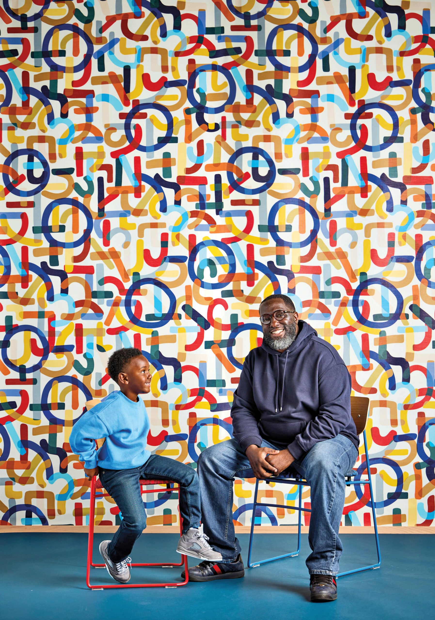 Michael Ford and his son, Mason, sit on chairs in front of a colorful, abstract-patterned wall, facing each other and smiling.