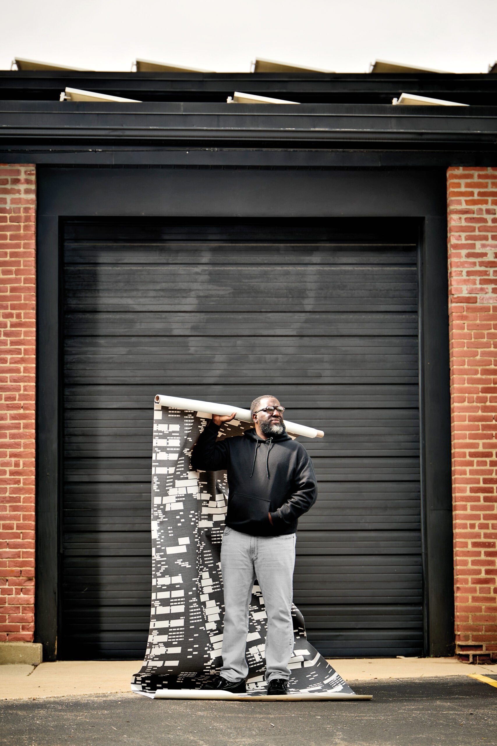 Michael Ford stands in front of a large black garage door, holding a rolled blueprint on his shoulder with more unrolled plans at his feet.