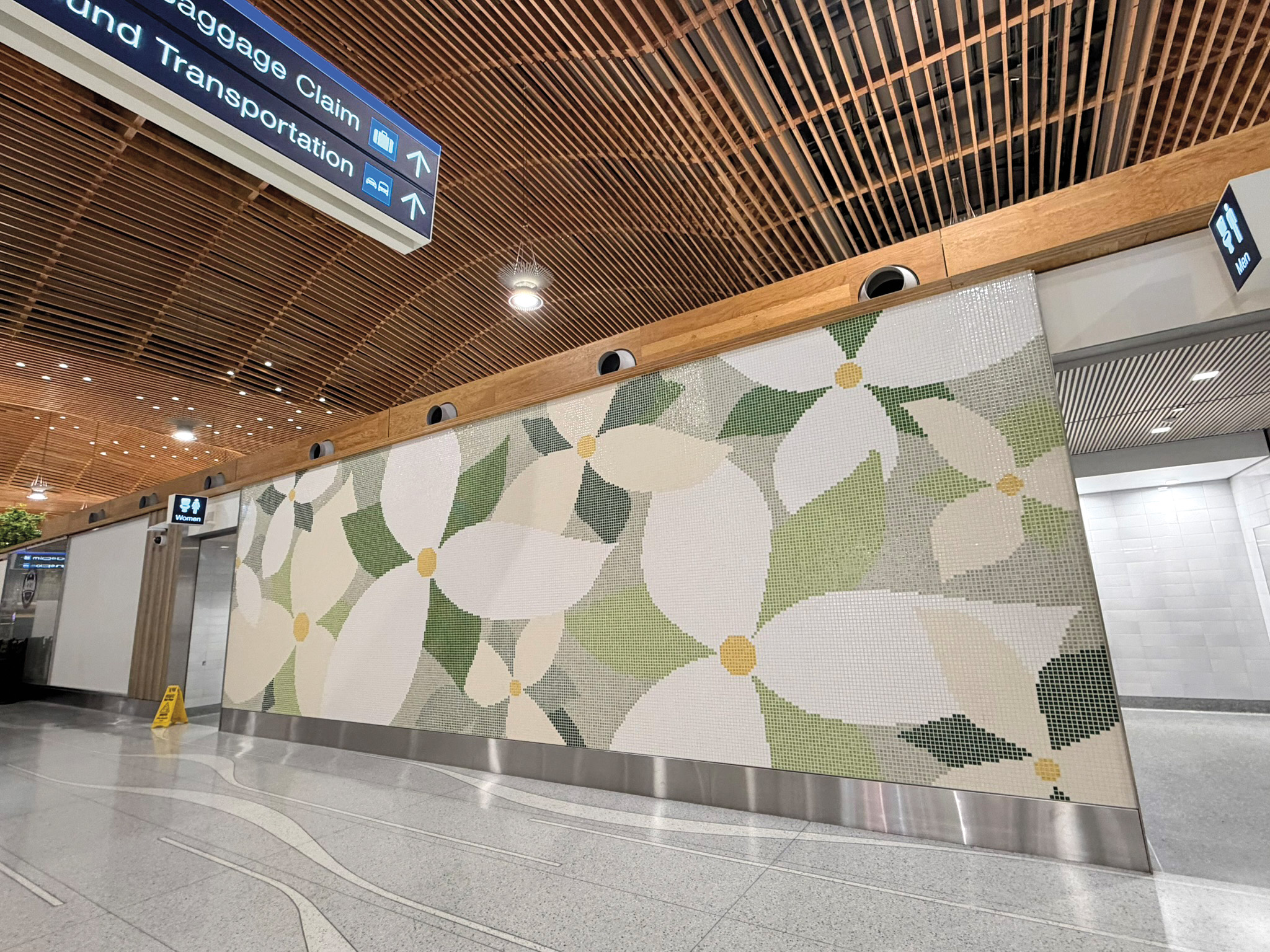 A large mosaic mural featuring white and green abstract floral patterns decorates a wall beneath ceiling signs in an airport terminal.