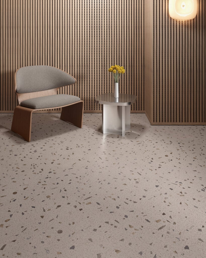 A modern room featuring terrazzo flooring, a wooden chair with gray cushion, a small round table displaying office products beside a yellow flower vase, and wooden slatted wall panels with a wall light.
