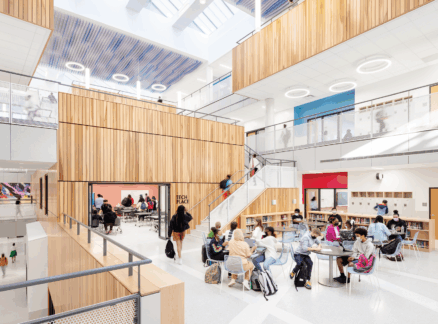 Students and adults gather and work in a bright, modern school atrium with wooden accents, large windows, and multiple seating areas—an inspiring space honored with planet positive awards.