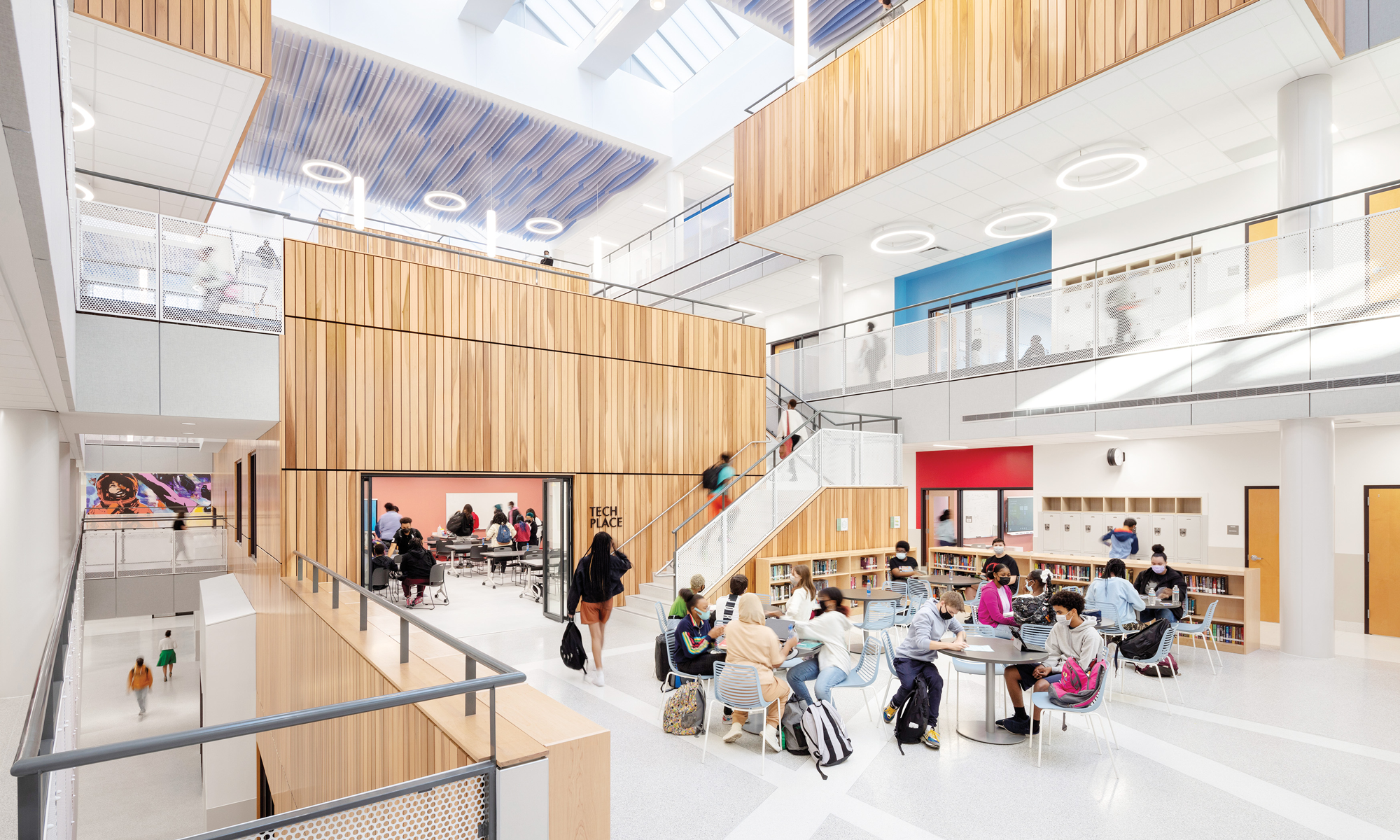 Students and adults gather and work in a bright, modern school atrium with wooden accents, large windows, and multiple seating areas—an inspiring space honored with planet positive awards.
