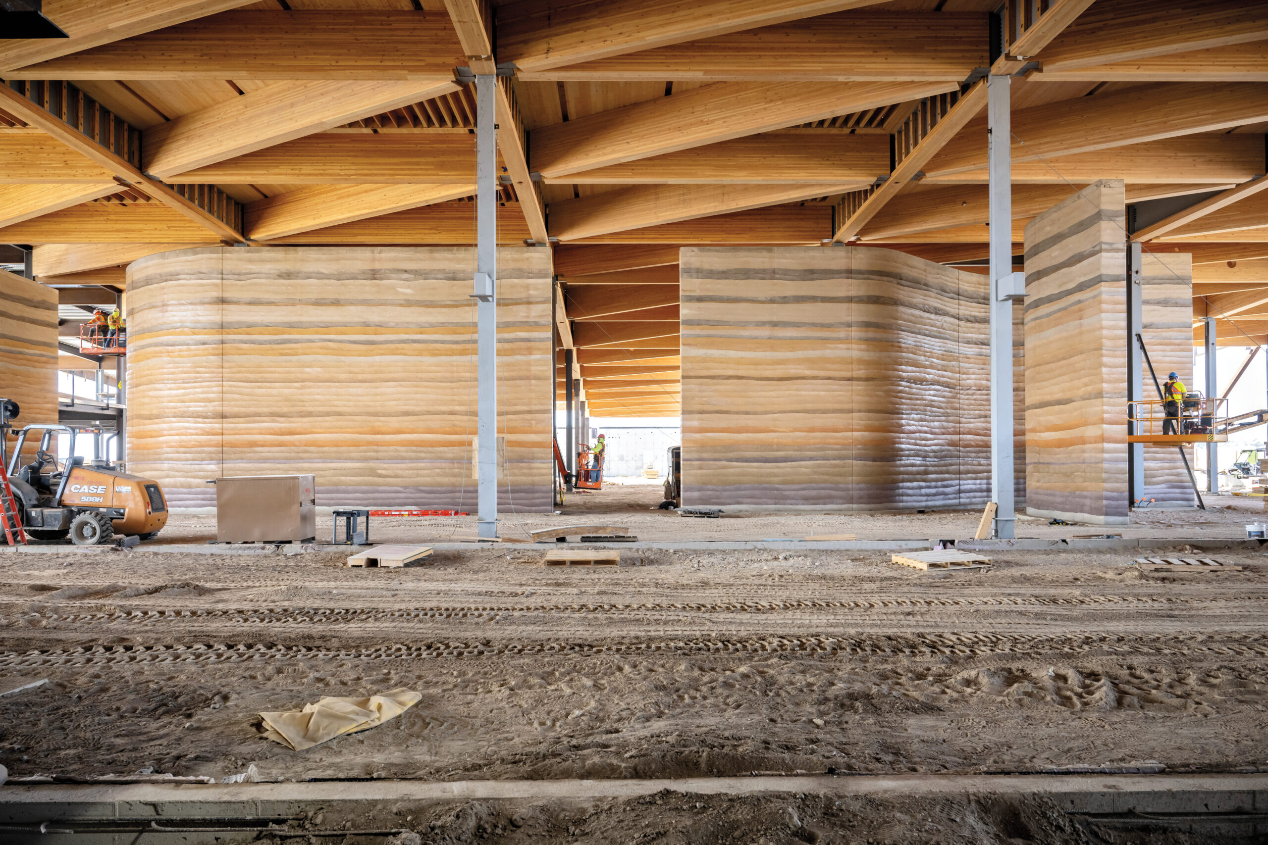 Interior view of a construction site featuring large rammed earth walls and exposed wooden beams, with construction equipment and workers visible.