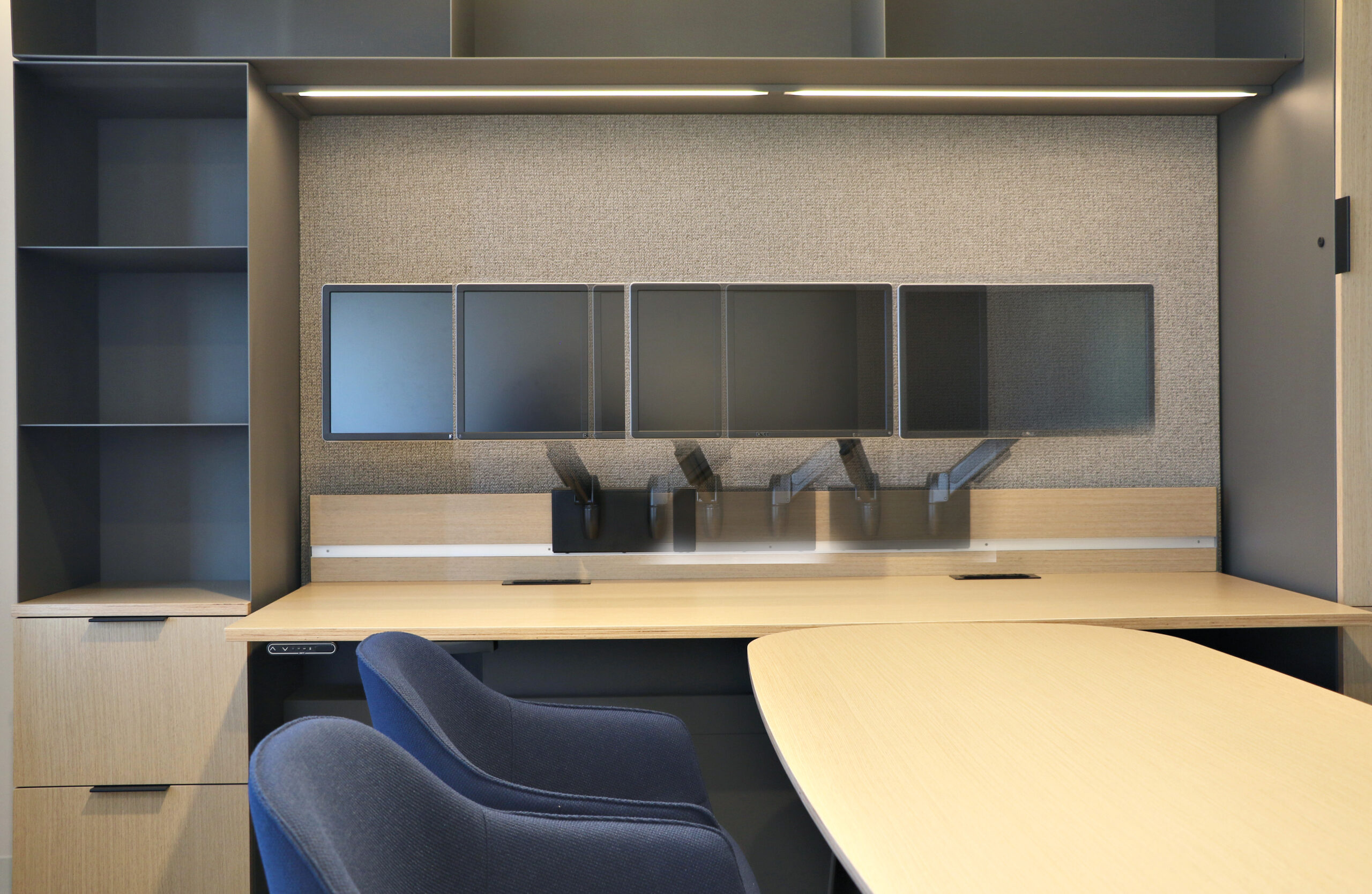 Modern office workspace with a light wood desk, blue chairs, empty shelving, and multiple wall-mounted monitor screens above the desk.
