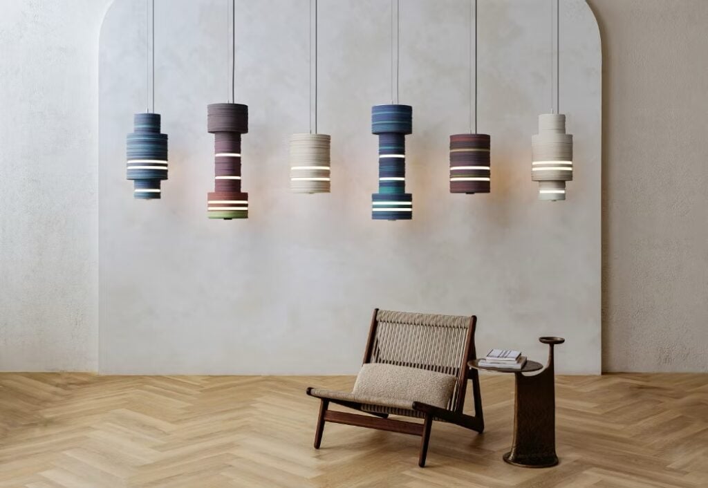 A modern room with six colorful pendant lights, sleek office products on a small side table, a wooden lounge chair with woven seat, and light herringbone wood flooring.