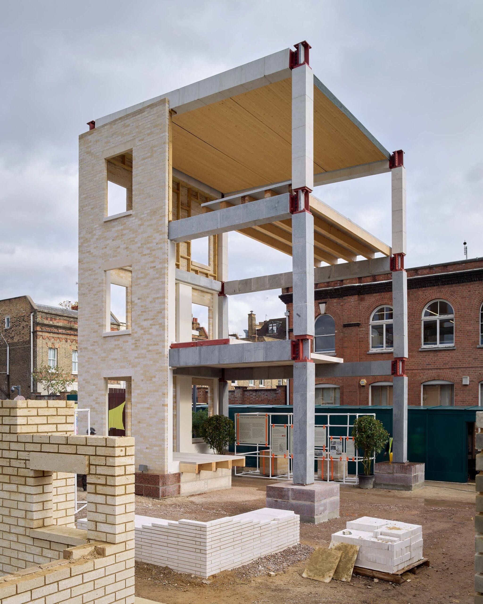 A partially constructed three-story building with exposed concrete, brick, and stone walls, open floors, and construction materials in the foreground on a cloudy day.