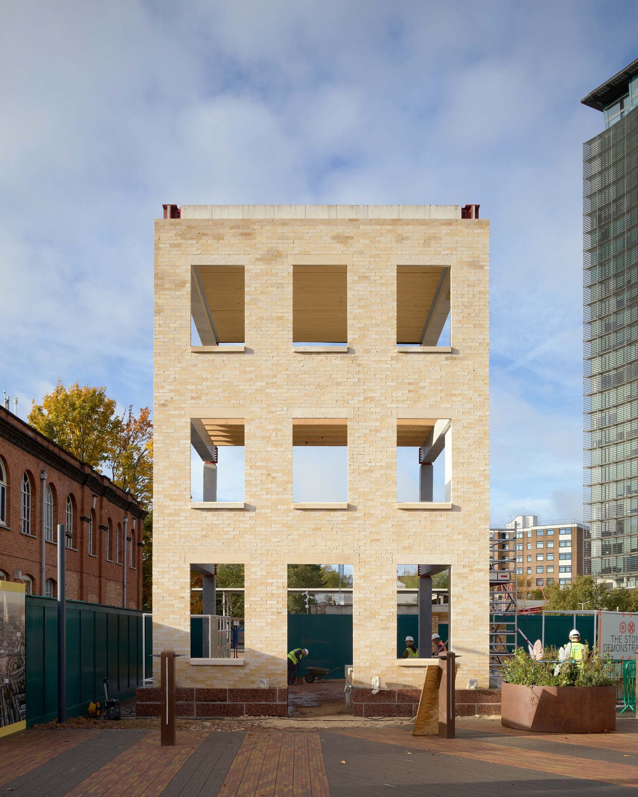 A rectangular three-story brick and stone building facade stands alone at a construction site, with workers and equipment visible around the base.