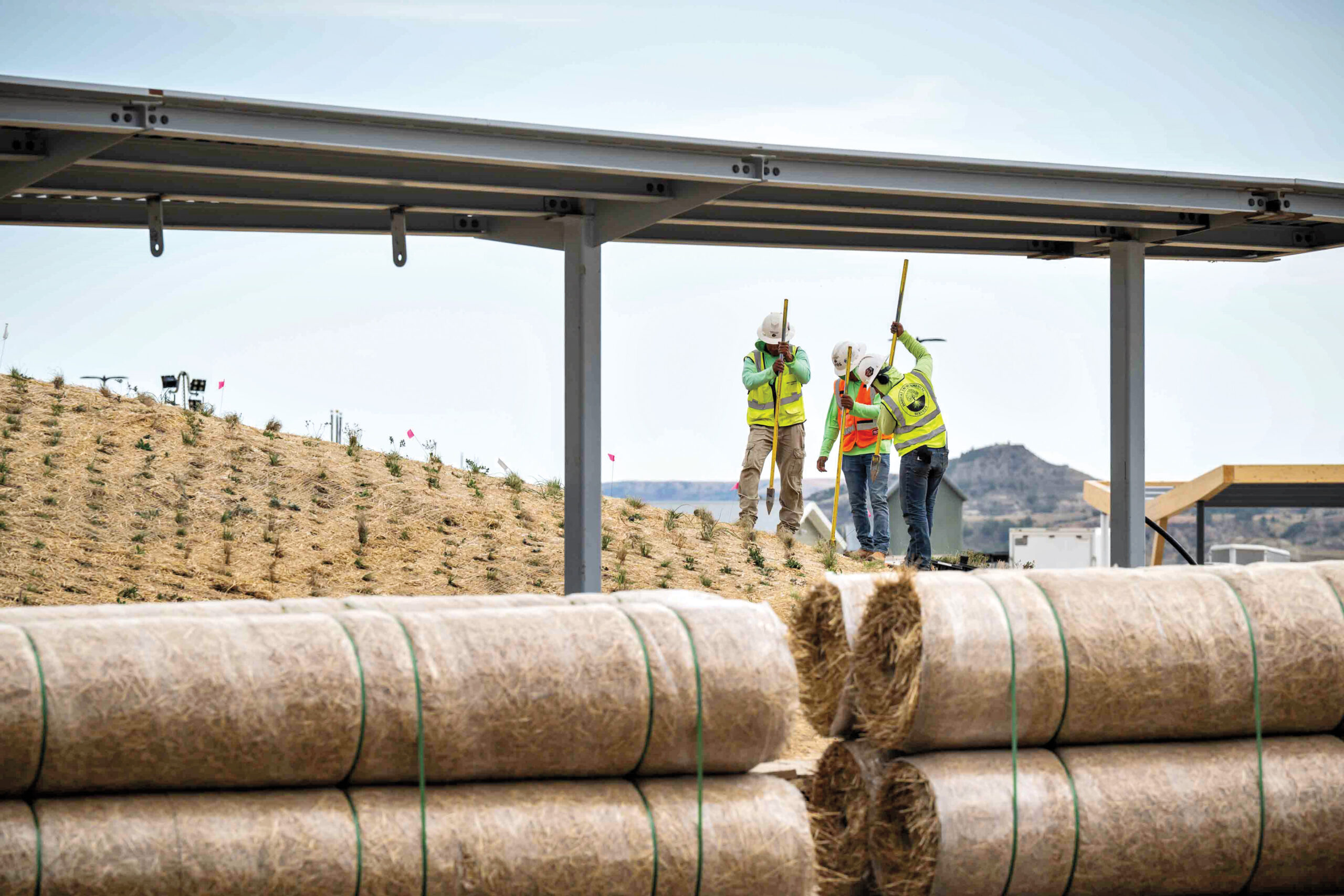 Three construction workers in safety gear work on a sloped, mulched landscape near large rolls of straw and a metal structure, with mountains visible in the background.