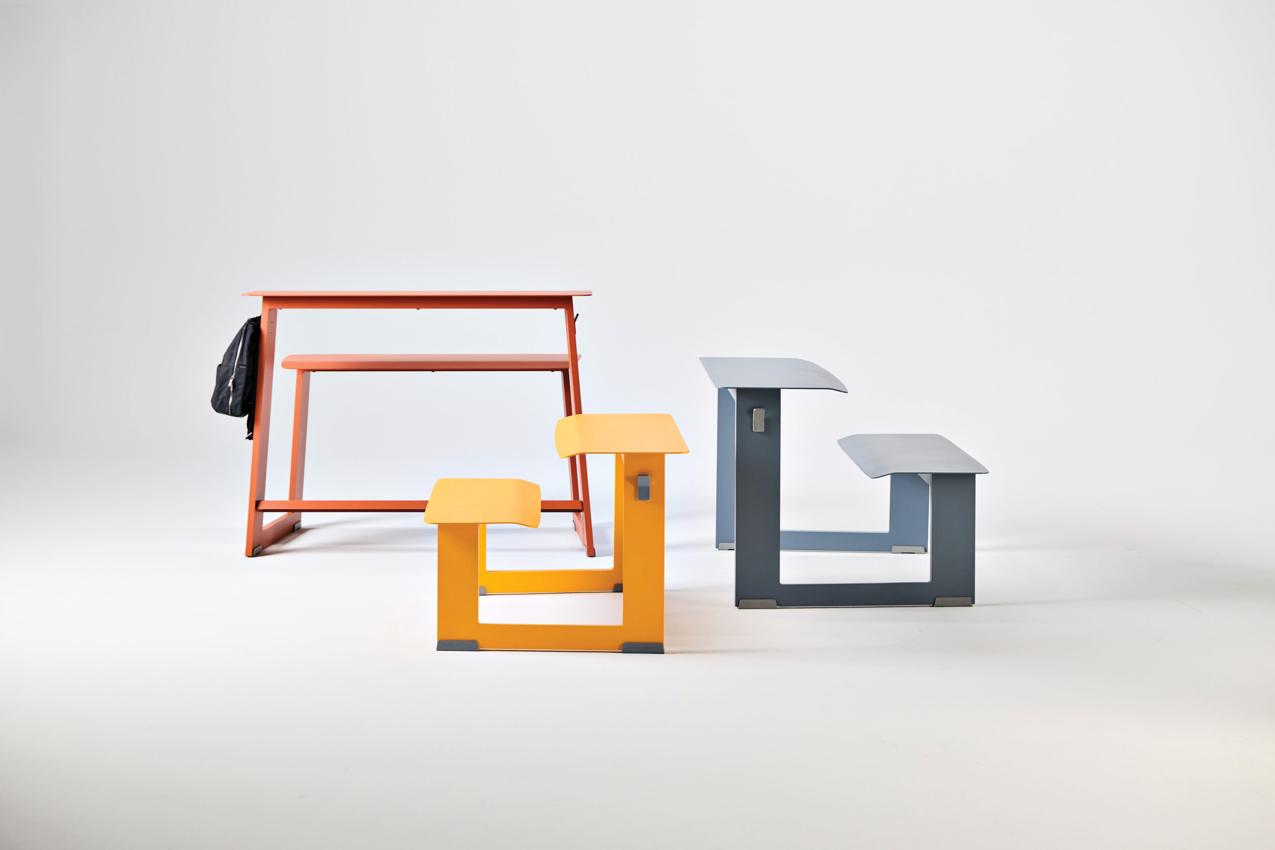 A red desk and matching bench are positioned behind two modern, L-shaped stools in yellow and gray, all set against a plain white background.