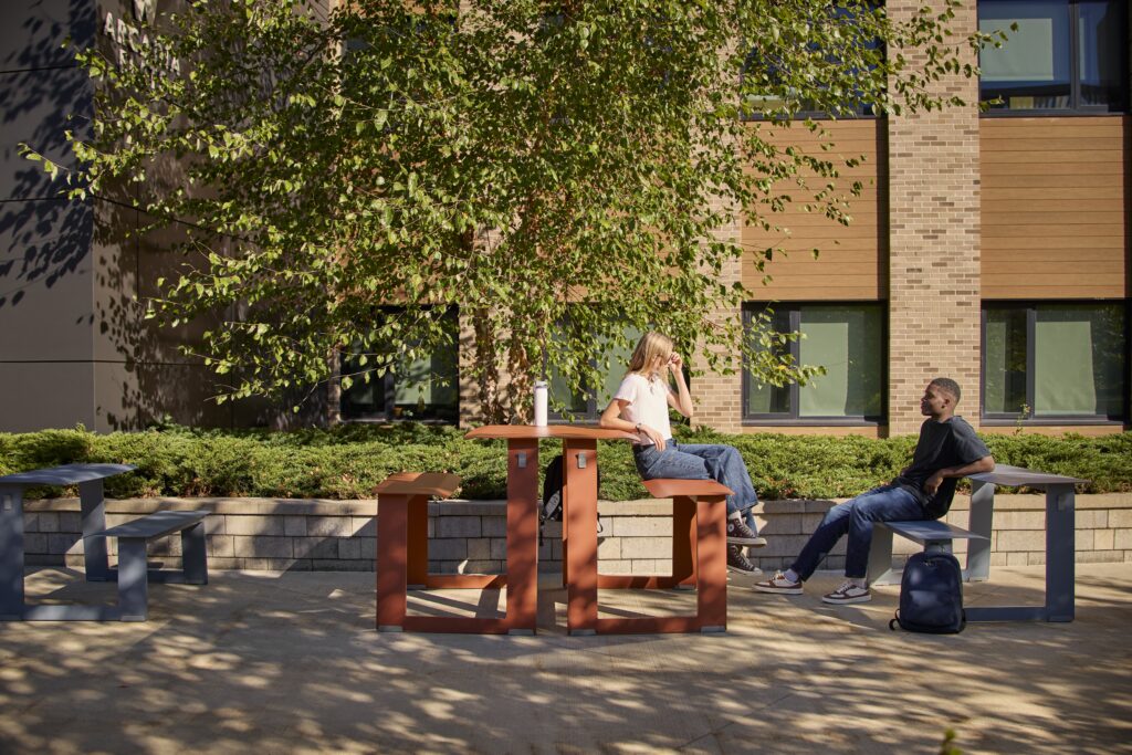 Two people sit and talk at an outdoor table in a courtyard, with trees, shrubs, and a brick building in the background.