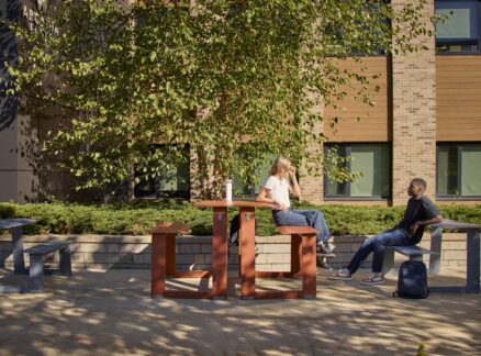 Two people sit and talk at an outdoor table in a courtyard, with trees, shrubs, and a brick building in the background.