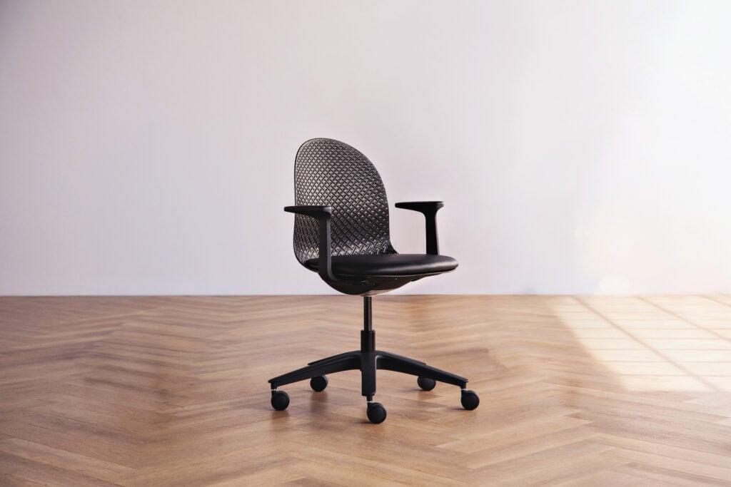 A black mesh office chair with armrests and wheels sits on a wooden herringbone floor against a plain light-colored wall, the perfect streamlined addition to any workspace admired by METROPOLISLikes.