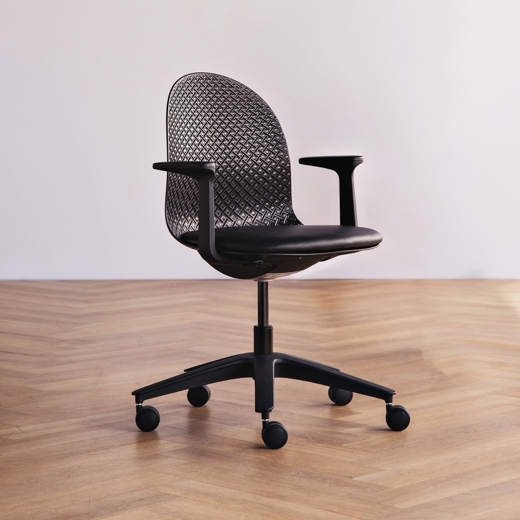 A black office chair with a mesh backrest, armrests, and five caster wheels sits on a wooden herringbone floor against a plain light-colored wall.