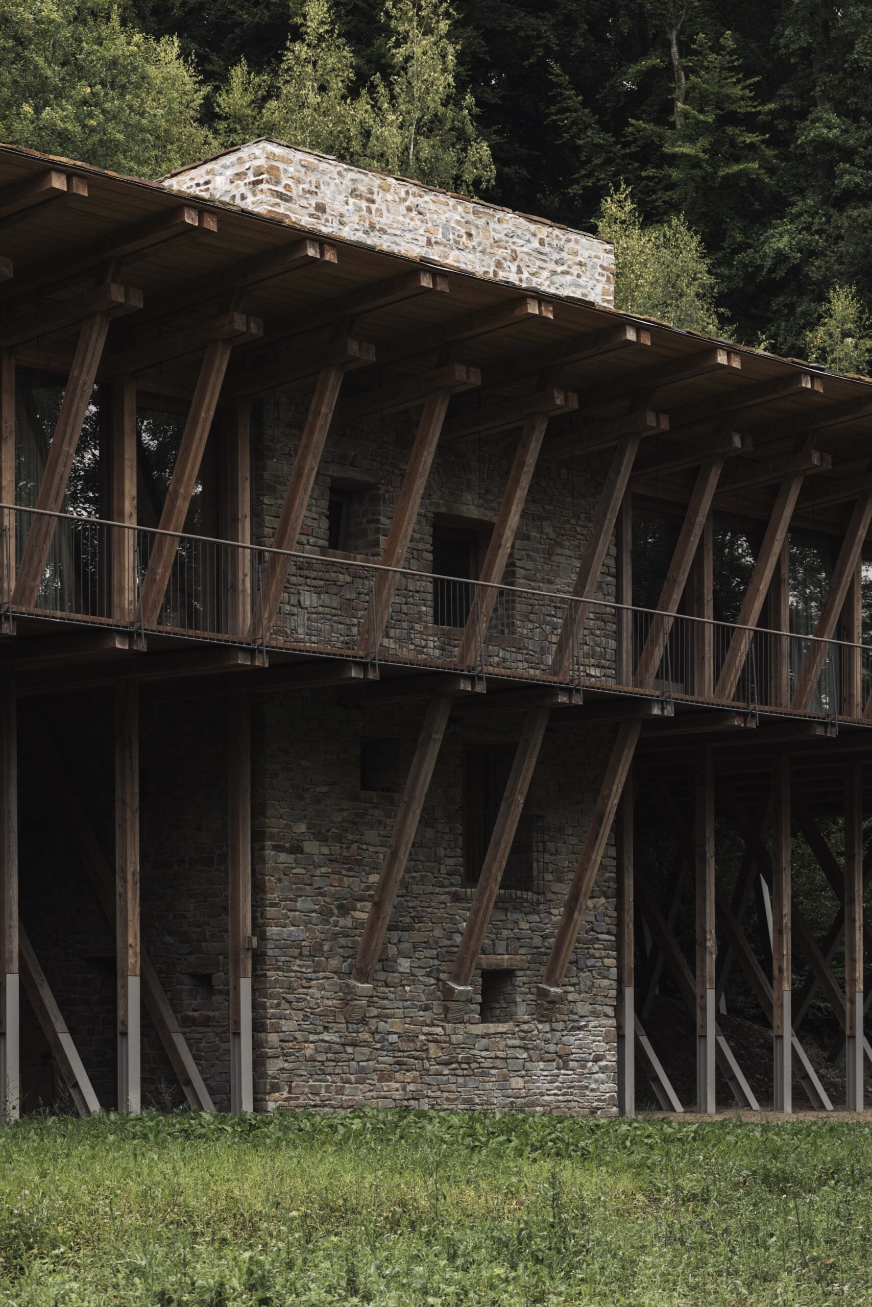 Two-story stone building with wooden beams, featuring an upper deck and surrounded by lush greenery and trees.