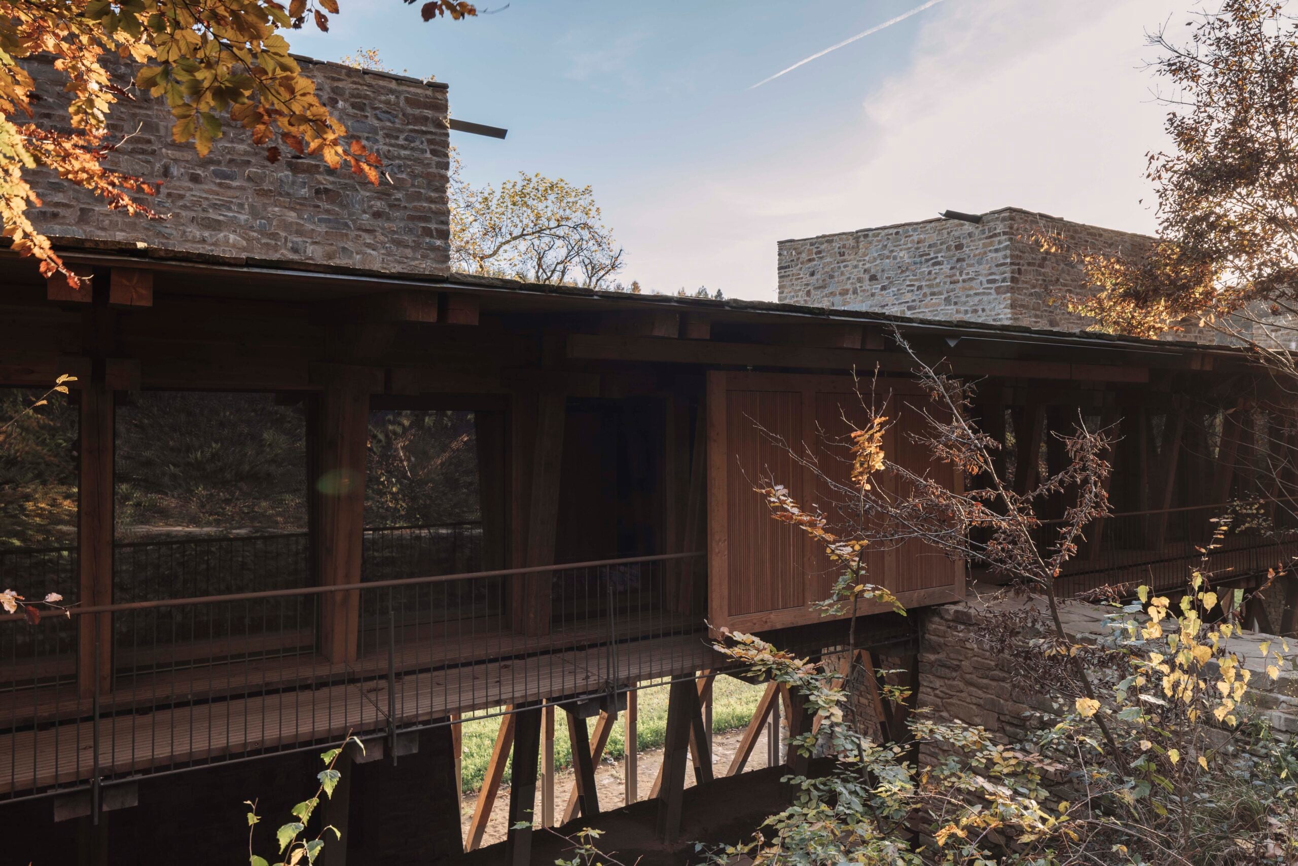 A modern stone building with elegant walls and a wooden bridge walkway, surrounded by autumn trees under a clear sky.