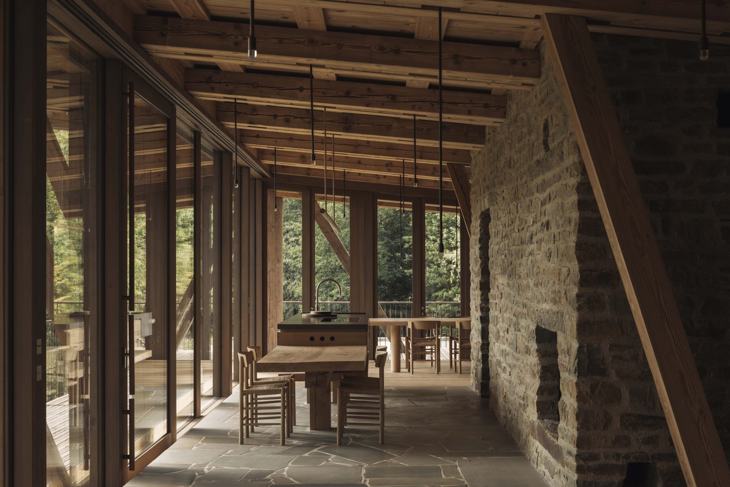 Modern dining area with exposed stone wall, wooden beams, large glass windows, and a long wooden table with chairs, overlooking a wooded outdoor view.