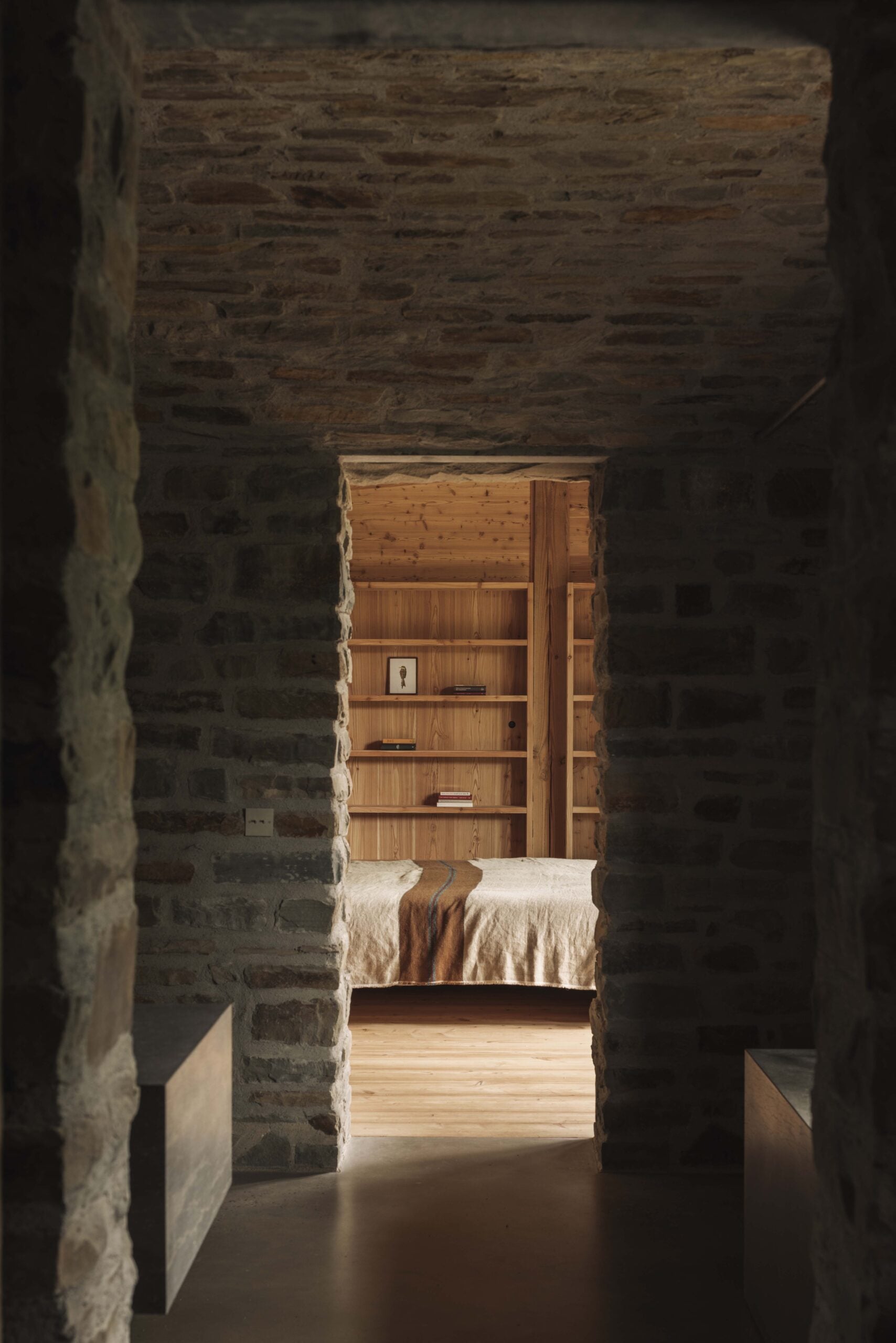 A stone-walled hallway leads to a softly lit bedroom with wooden walls, a neatly made bed, and built-in stone shelves.