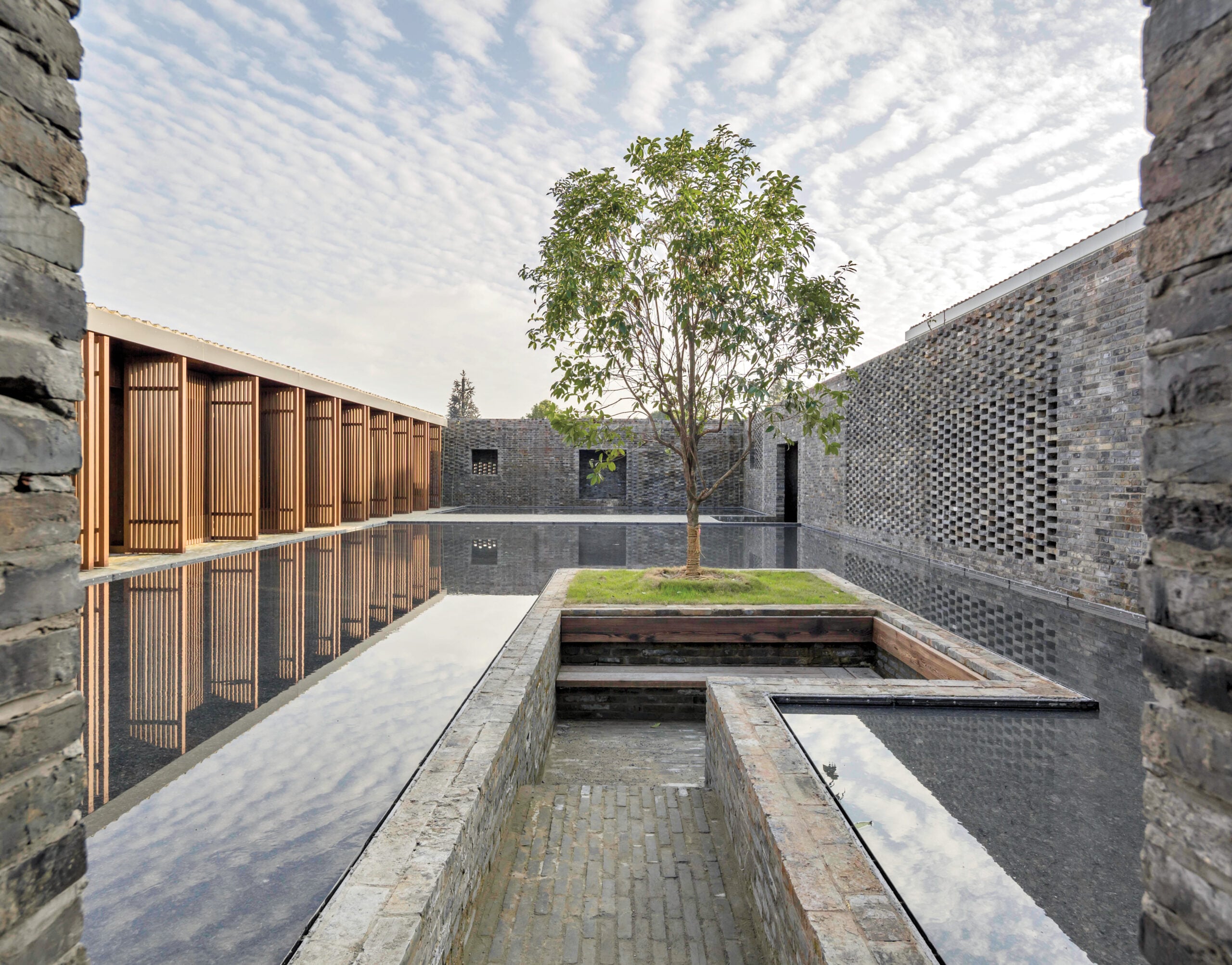 A courtyard with a single tree, surrounded by reflective water and bordered by stone and wood walls under a partly cloudy sky.