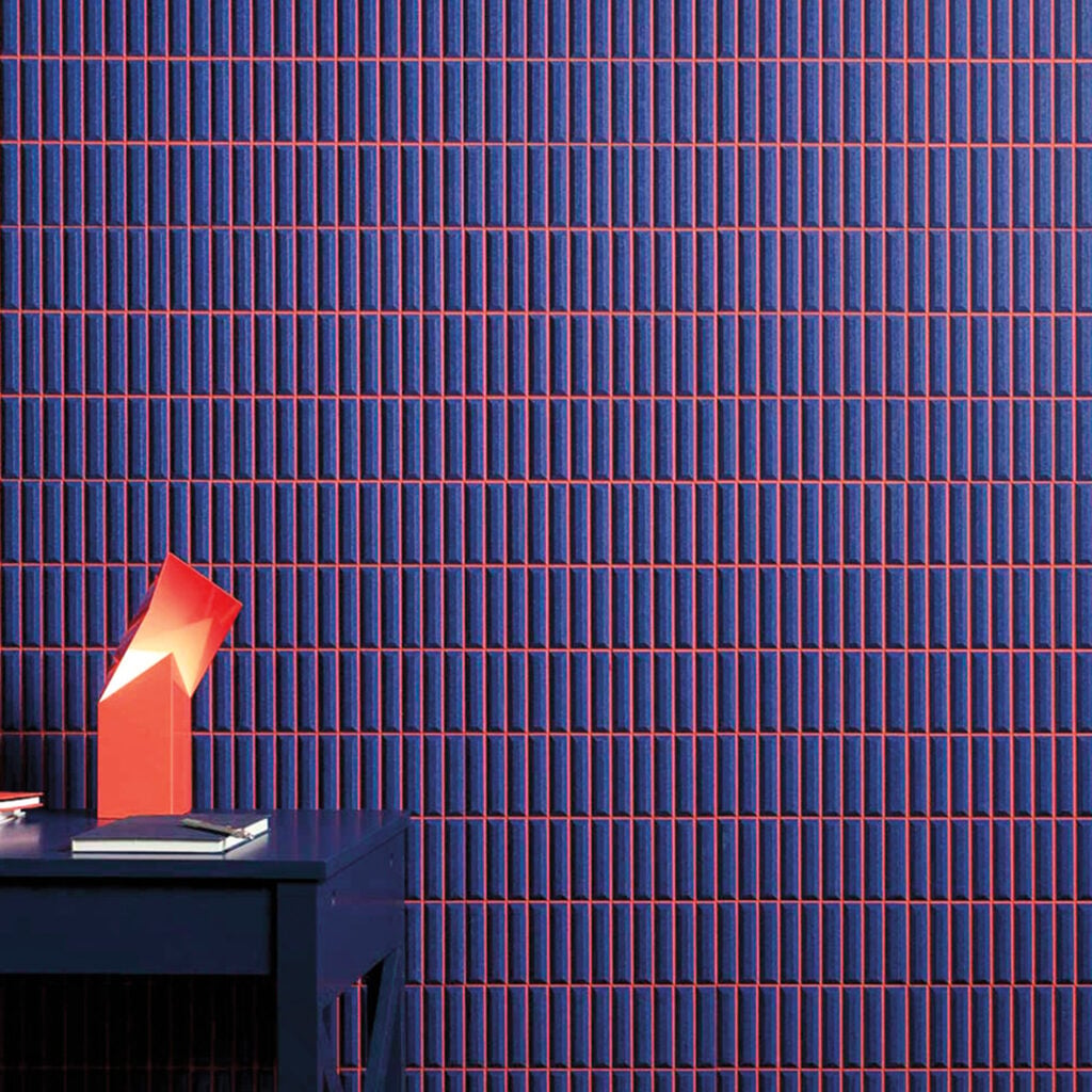 A blue desk with stacked books and a geometric red object sits in front of a wall covered in a navy and red linear grid-patterned wallpaper.