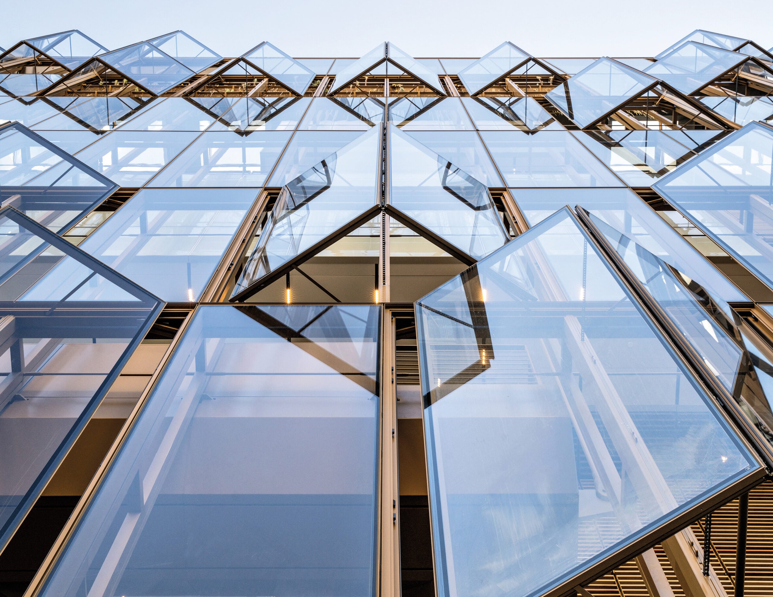 Craft in Architecture: A modern building facade with multiple glass windows open outward in a geometric pattern, reflecting the clear sky above.