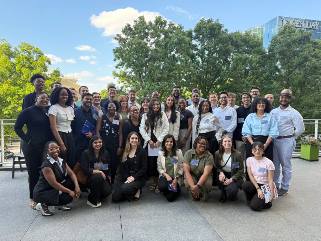 A diverse group of people pose and smile for an Arch30 group photo outdoors on a sunny day, with trees and a modern building in the background.