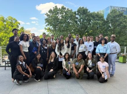 A diverse group of people pose and smile for an Arch30 group photo outdoors on a sunny day, with trees and a modern building in the background.
