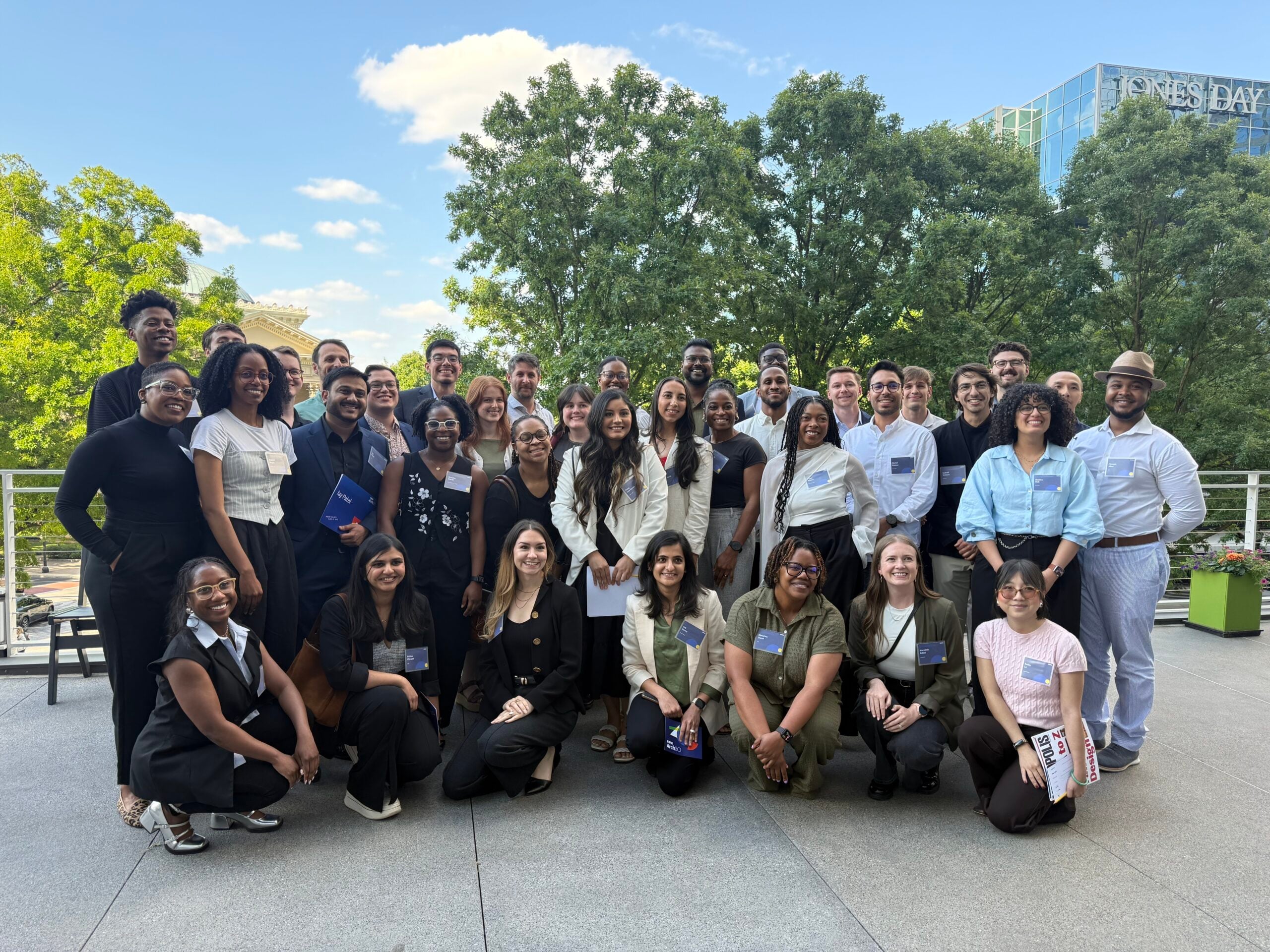A diverse group of people pose and smile for an Arch30 group photo outdoors on a sunny day, with trees and a modern building in the background.