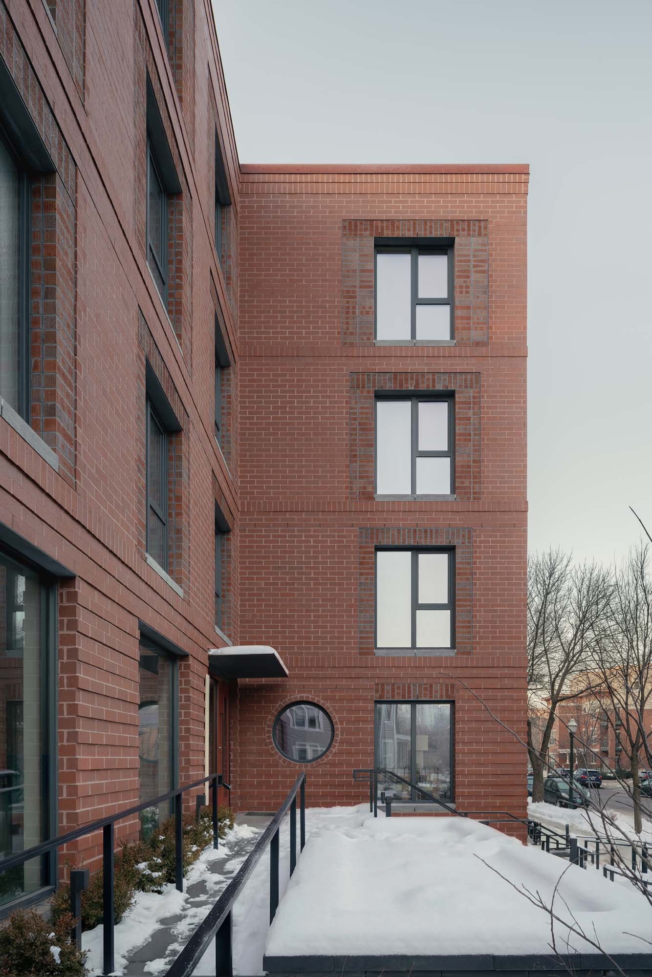 A modern brick apartment building with large rectangular windows and a circular window by the entrance. Snow covers the ground and nearby ledges; bare trees are visible in the background.