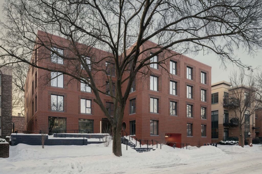 A modern, four-story brick apartment building with large windows, set behind a leafless tree and surrounded by snow on the ground and sidewalks.